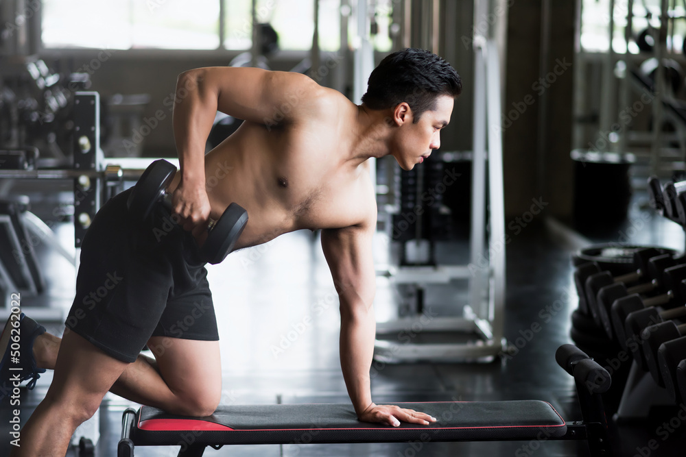 Fit Asian young man doing one-arm dumbbell on bench in gym
