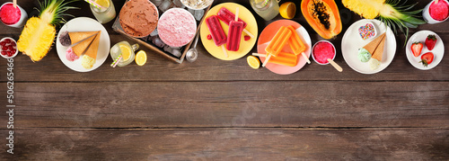 Cool summer foods top border. Selection of ice cream, popsicles and fruit. Above view over a dark wood banner background.