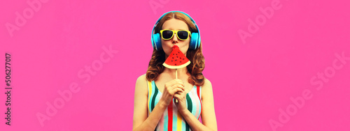 Summer portrait of stylish woman in headphones listening to music blowing her lips with juicy lollipop or ice cream shaped slice watermelon on pink background  blank copy space for advertising text