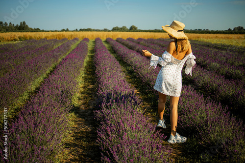 Back view of a pretty young woman in white dress in straw hat running walking at the sage flower field. Beautiful girl enjoying a field of flowers, relaxing outdoors, having fun, harmony concept.
