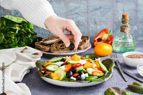 A woman's hand puts an olive in a freshly prepared Greek salad on a plate on the table.