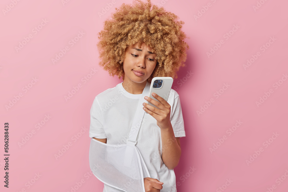 Indoor shot of serious displeased woman with curly hair wears sling on broken arm suffers from pain after accident searches advice how to cure in internet focused in smartphone screen isolated on pink Indoor shot of serious displeased woman with curly hair wears sling on broken arm suffers from pain after accident searches advice how to cure in internet focused in smartphone screen isolated on pink