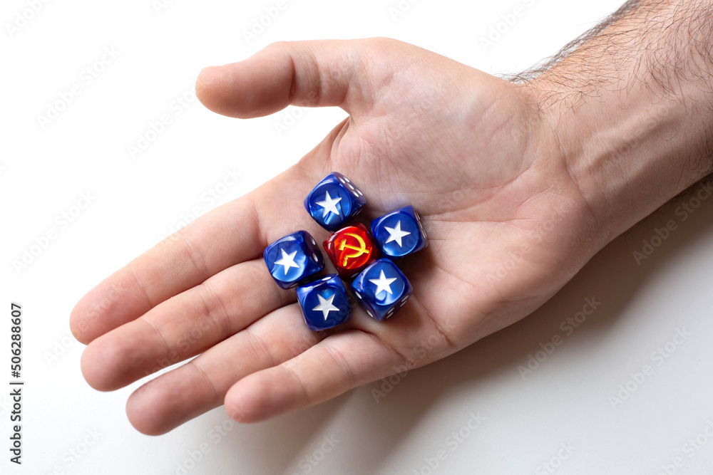 One dice with the symbol hammer and sickle surrounded by five blue cubes with the symbol usa star are on a man's palm. The concept of the ideology of communism and capitalism. One dice with the symbol hammer and sickle surrounded by five blue cubes with the symbol usa star are on a man's palm. The concept of the ideology of communism and capitalism.