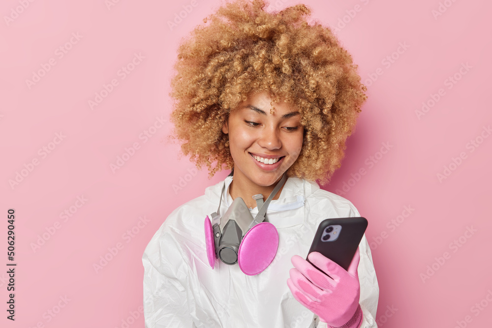 Positive woman dressed in protective suit and gloves looks at smartphone screen fights against air pollution isolated over pink background. Female ecologist explores problems on environment. Positive woman dressed in protective suit and gloves looks at smartphone screen fights against air pollution isolated over pink background. Female ecologist explores problems on environment.