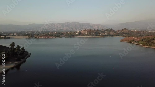 aerial footage of a vast blue rippling lake at sunset surrounded by lush green trees, grass and plants with majestic mountain ranges and blue sky at Puddingstone Lake in San Dimas California USA photo