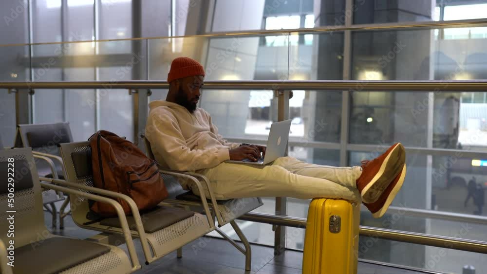 Focused young African American man sitting in hall of airport terminal working on laptop. Concentrated freelancer black male online working on computer and waiting landing on board. Business travel. Focused young African American man sitting in hall of airport terminal working on laptop. Concentrated freelancer black male online working on computer and waiting landing on board. Business travel.