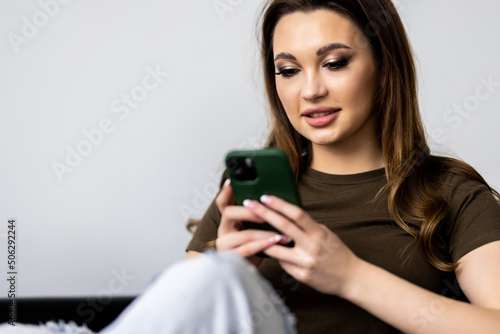 Smiling young woman using phone, sitting on couch at home, looking at smartphone screen