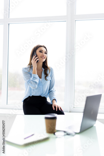 Good business talk. Cheerful young beautiful woman in glasses talking on mobile phone and using laptop with smile while sitting at her working place © F8  \ Suport Ukraine