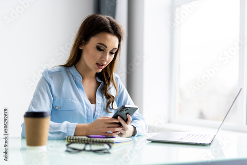 Freelance woman in glasses with mobile phone typing at laptop and working from home office with plants. Happy girl on workplace at the desk. Distance learning online education and work.