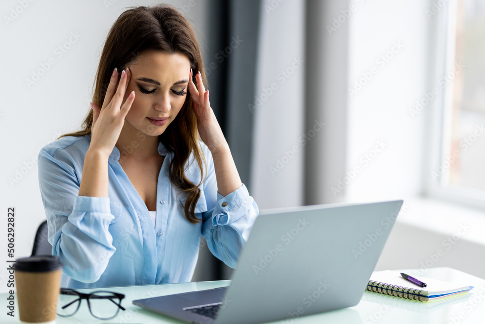 Young attractive woman at modern office desk, working on laptop, massaging temples to forget about constant headaches, noisy loud office giving a migraine, relieving stress, chronic pain, help soothe Young attractive woman at modern office desk, working on laptop, massaging temples to forget about constant headaches, noisy loud office giving a migraine, relieving stress, chronic pain, help soothe