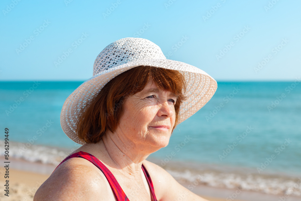 An elderly woman in a hat on the seashore on a sunny day, looks away and smiles, close-up. Summer pastime in retirement