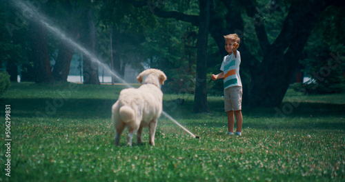 Little boy play water sprinkler on green field with playful golden retriever 