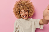 Positive young European woman with blonde curly hair keeps arm outstreched being in good mood makes photo with unrecognizable device dressed in casual t shirt isolated over pink studio background.