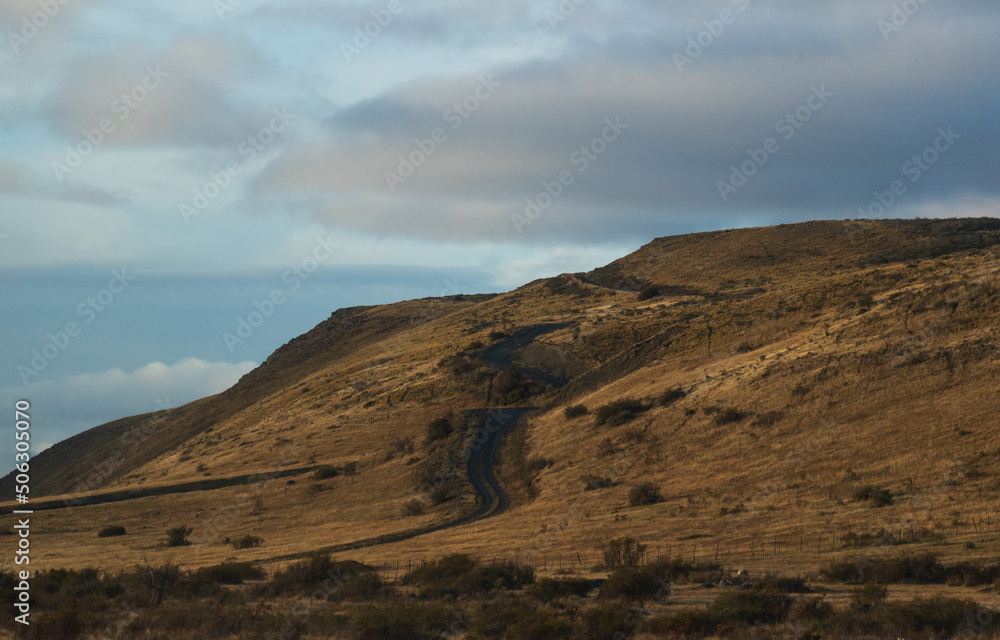 carretera alternativa de tierra y grava atravesando entre cerro y colinas de colores cafés otoñales y lluvia, con nubes nimbostratos , camino entre cerro