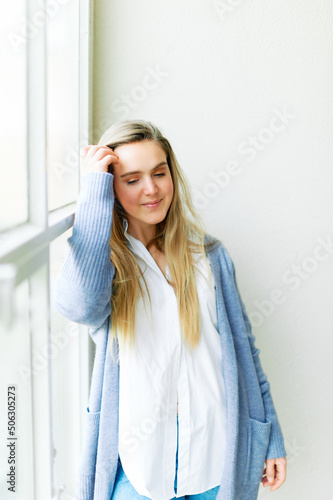 Portrait of young pretty 20 year old girl posing next to big window, wearing white shirt and blue cardigan, looking down Free Download Portrait of young pretty 20 year old girl posing next to big window, wearing white shirt and blue cardigan, looking down - GFXLoad