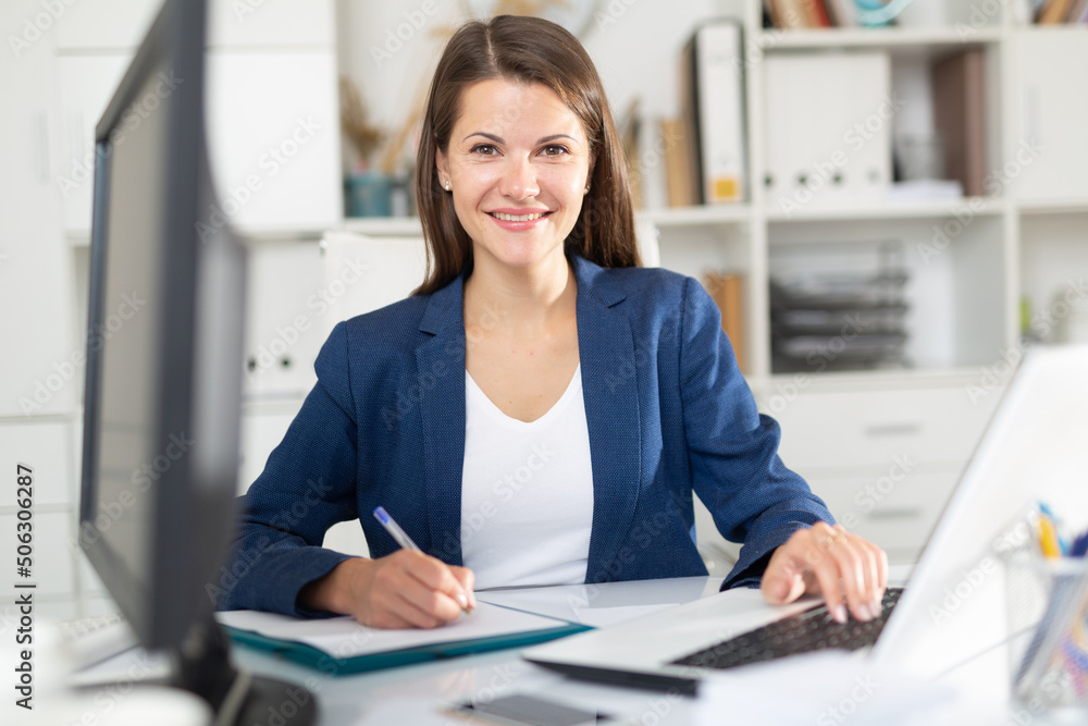 Portrait of confident smiling female office employee during daily work with laptop and documents - obrazy, fototapety, plakaty 