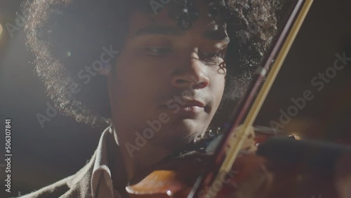 Handheld camera shot of young African American violin player performing music on stage in spotlight