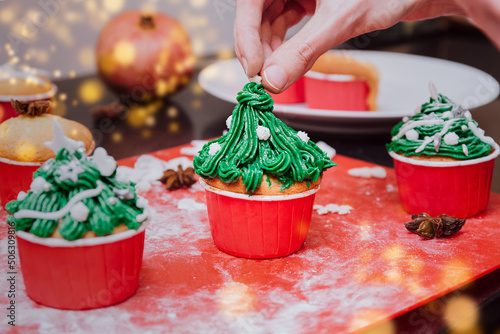 Christmas tree shaped cupcakes with cream frosting on christmas lights and festive background. Man hands Cooking Christmas cupcakes and decorate cupcakes with cream, Christmas treatment for family