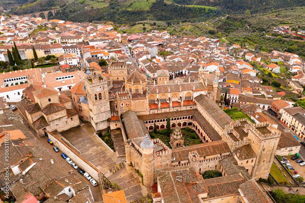 Gothic-Mudejar style building of Royal Monastery of Saint Mary in Spanish town of Guadalupe, located in green valley of province of Caceres overlooking brownish roofs of houses, as seen from drone..