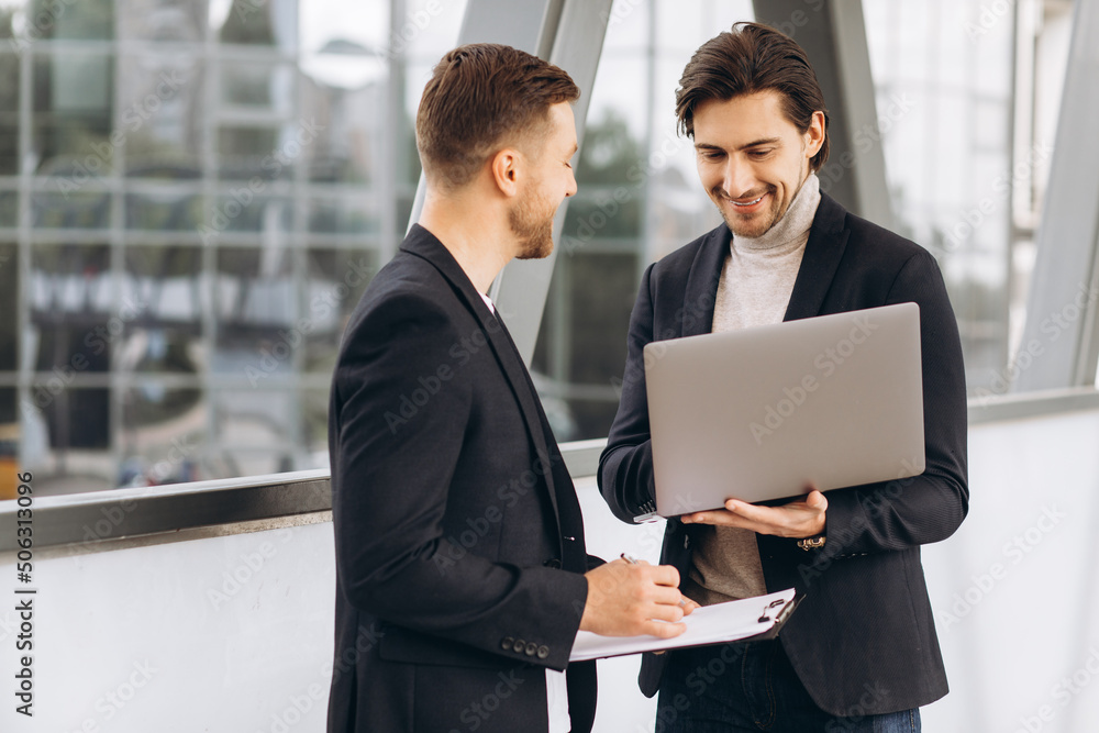 Two handsome young male businessmen in suits hold a laptop and a folder with documents and discuss a working project or have a video call against the background of urban office buildings. Two handsome young male businessmen in suits hold a laptop and a folder with documents and discuss a working project or have a video call against the background of urban office buildings.