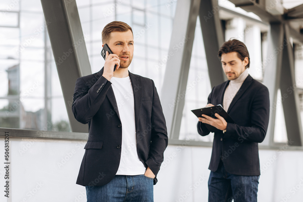 Two modern businessman with phone and documents folder discuss something and walk across the bridge against the background of urban offices and buildings Two modern businessman with phone and documents folder discuss something and walk across the bridge against the background of urban offices and buildings