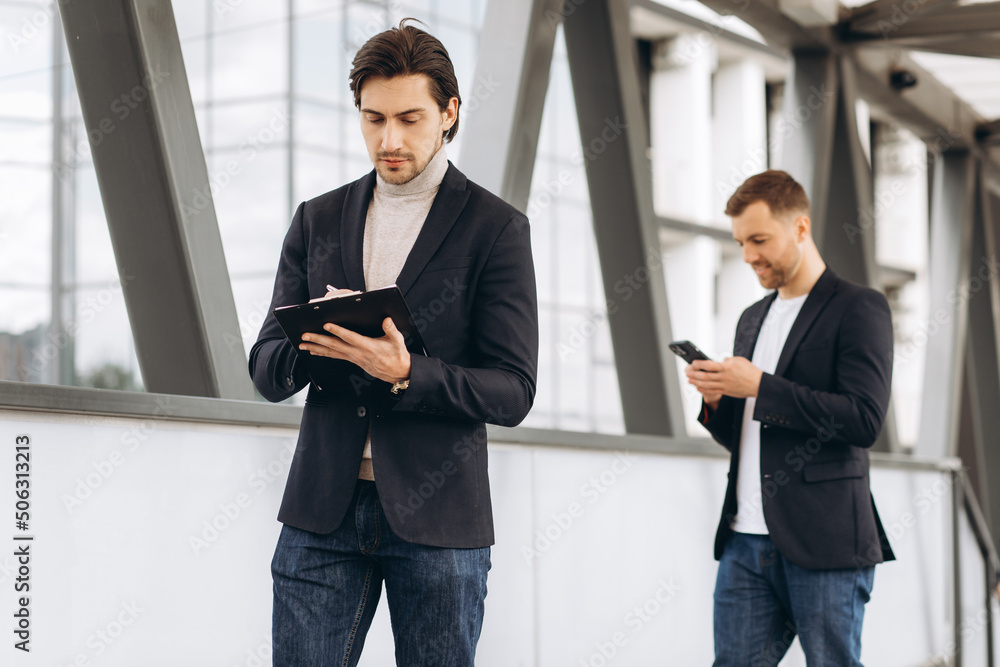 Modern and handsome businessman with a folder with documents writes something on the background of city offices and buildings Modern and handsome businessman with a folder with documents writes something on the background of city offices and buildings