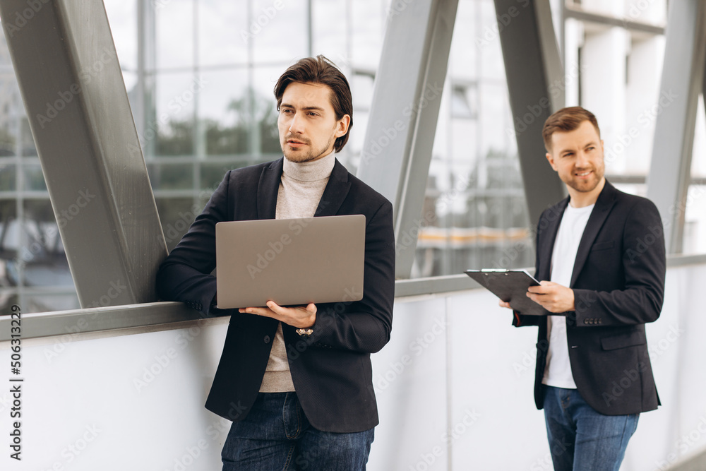 Two modern happy businessmen with laptop and document folder discussing something against the background of urban offices and buildings Two modern happy businessmen with laptop and document folder discussing something against the background of urban offices and buildings