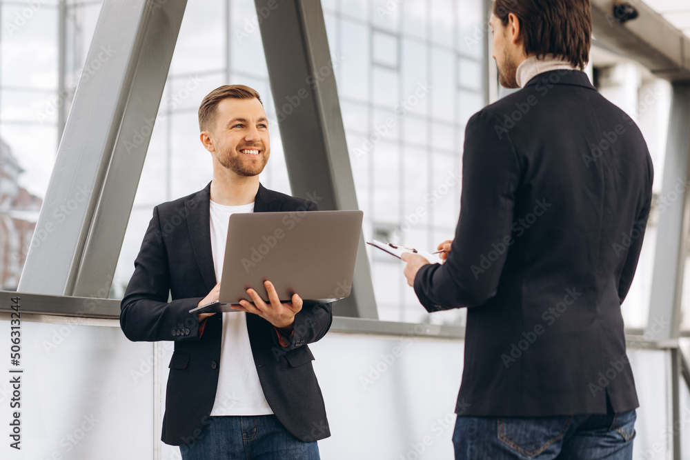 Two modern happy businessmen with laptop and document folder discussing something against the background of urban offices and buildings Two modern happy businessmen with laptop and document folder discussing something against the background of urban offices and buildings