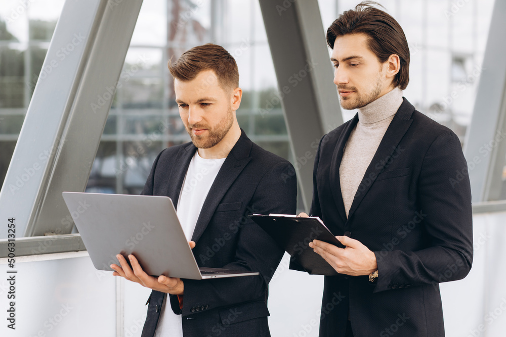 Two modern happy businessmen with laptop and document folder discussing something against the background of urban offices and buildings Two modern happy businessmen with laptop and document folder discussing something against the background of urban offices and buildings