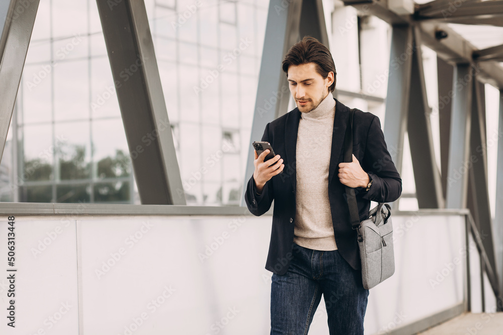Portrait of handsome modern male businessman in suit walking smiling and reading or writing message on phone on background of urban buildings and offices. Portrait of handsome modern male businessman in suit walking smiling and reading or writing message on phone on background of urban buildings and offices.