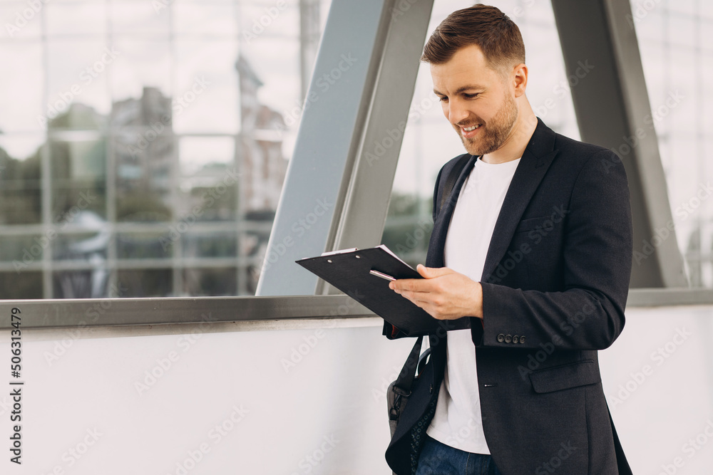 Portrait of handsome modern male businessman holding folder with documents on background of urban buildings and offices. Portrait of handsome modern male businessman holding folder with documents on background of urban buildings and offices.