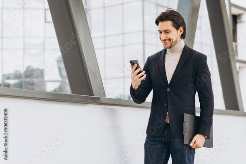 Portrait of handsome modern male businessman in suit holding folder with documents and talking on the phone against the background of urban buildings and offices.
