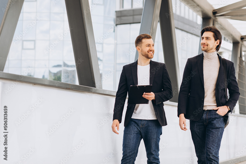 Two modern happy businessmen walking across a city bridge discussing something against the background of urban offices and buildings Two modern happy businessmen walking across a city bridge discussing something against the background of urban offices and buildings