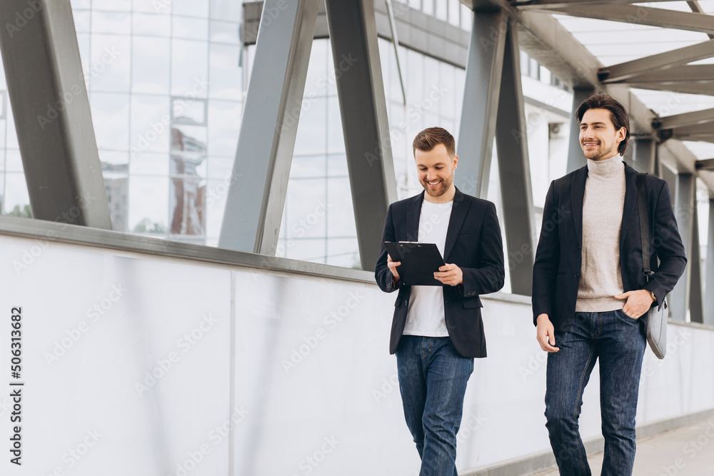 Two modern happy businessmen walking across a city bridge discussing something against the background of urban offices and buildings Two modern happy businessmen walking across a city bridge discussing something against the background of urban offices and buildings