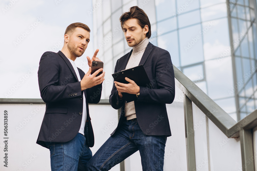 Two modern happy businessmen with phone and folder of documents discussing something against the background of urban offices and buildings Two modern happy businessmen with phone and folder of documents discussing something against the background of urban offices and buildings