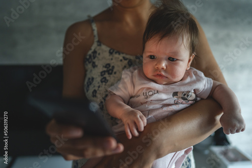 Adult woman mother holding three months old baby while using looking mobile phone for internet browsing or social network texting in room at home domestic life motherhood and parenting busy concept