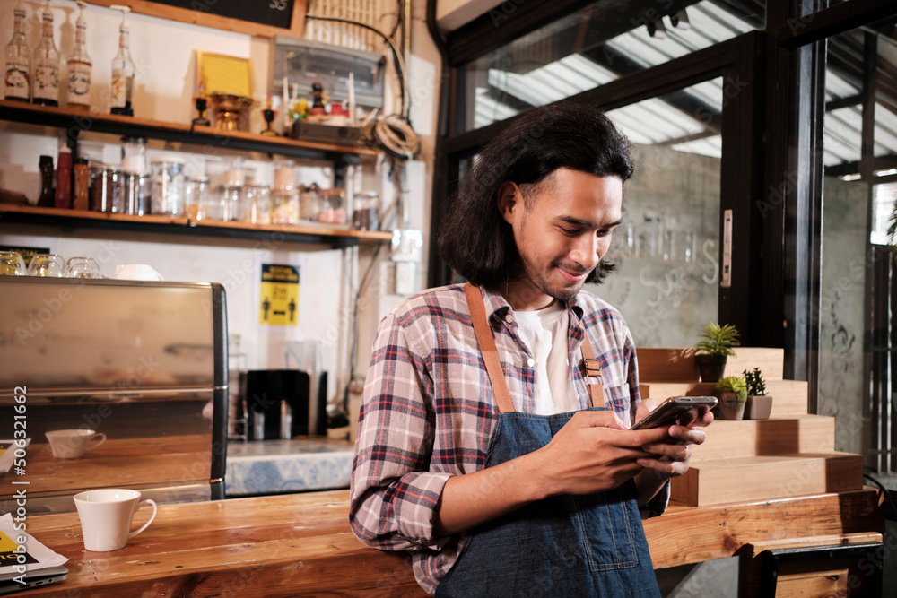 Long hair handsome Asian male startup barista with an apron stands at a casual counter bar cafe, chats about online orders in mobile phone application, and coffee shop service jobs entrepreneur. Long hair handsome Asian male startup barista with an apron stands at a casual counter bar cafe, chats about online orders in mobile phone application, and coffee shop service jobs entrepreneur.