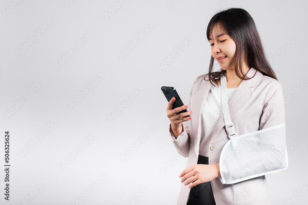 Woman confident smiling broken arm after accident and wear arm splint for treatment and hold smartphone, happy Asian female sling supported hand isolated on white background, social media, copy space Woman confident smiling broken arm after accident and wear arm splint for treatment and hold smartphone, happy Asian female sling supported hand isolated on white background, social media, copy space