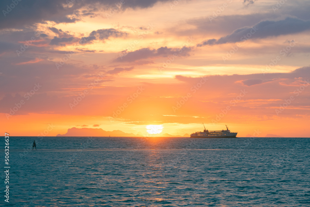 The sun setting in the sea against the backdrop of the islands and the sailing ferry.  Colored skies over the sea.  A man stands on a sandy spit in the sea and enjoy the sunset.