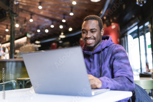 Positive African American male freelancer sitting at table and typing on netbook while working remotely in modern cafe