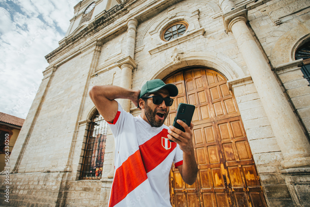 Hincha peruano de futbol celebrando un gol mirando su telefono en un parque de peru. Concepto de Personas y deportes. - obrazy, fototapety, plakaty 