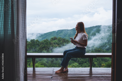 A young asian woman using mobile phone while sitting on balcony with a beautiful nature view on foggy day © Farknot Architect