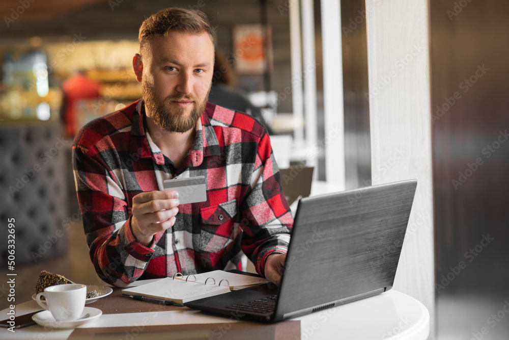 Portrait of amazing bearded young man sitting in cafe using laptop computer and holding debit card. Smiling and shopping online with credit card. Portrait of amazing bearded young man sitting in cafe using laptop computer and holding debit card. Smiling and shopping online with credit card.