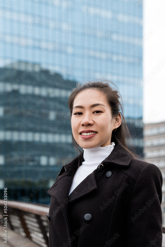 Positive Chinese female manager in black coat smiling and looking at camera while standing on bridge near glass skyscraper in downtown Positive Chinese female manager in black coat smiling and looking at camera while standing on bridge near glass skyscraper in downtown