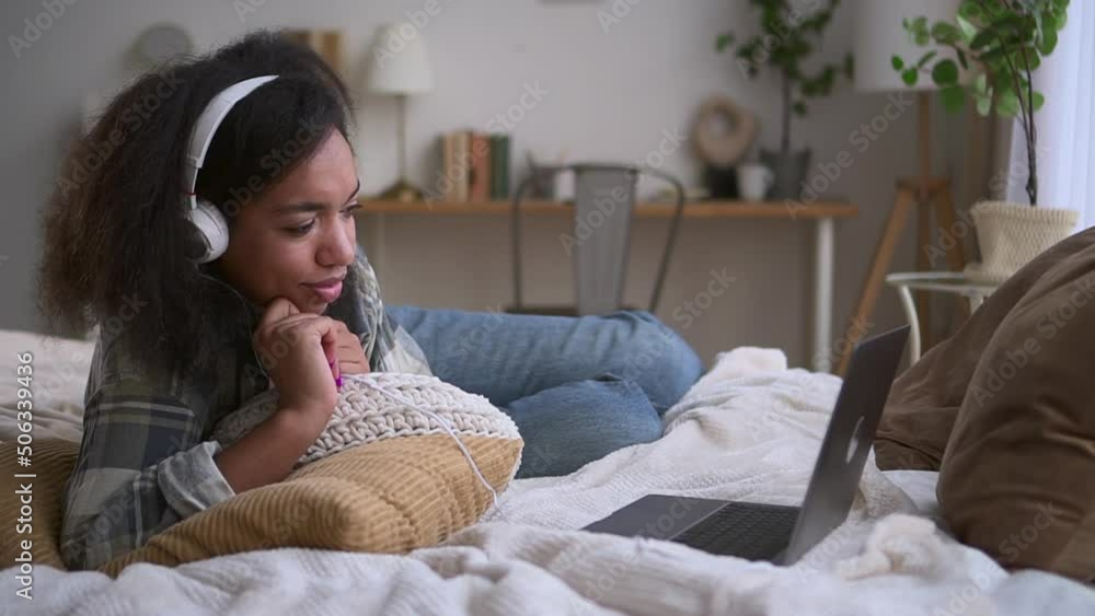 Young beautiful woman is chatting with laptop and lying on bed in home bedroom spbd. Close view of american african person looks at computer screen and talks with smile, rests and lies in cozy room Young beautiful woman is chatting with laptop and lying on bed in home bedroom spbd. Close view of american african person looks at computer screen and talks with smile, rests and lies in cozy room