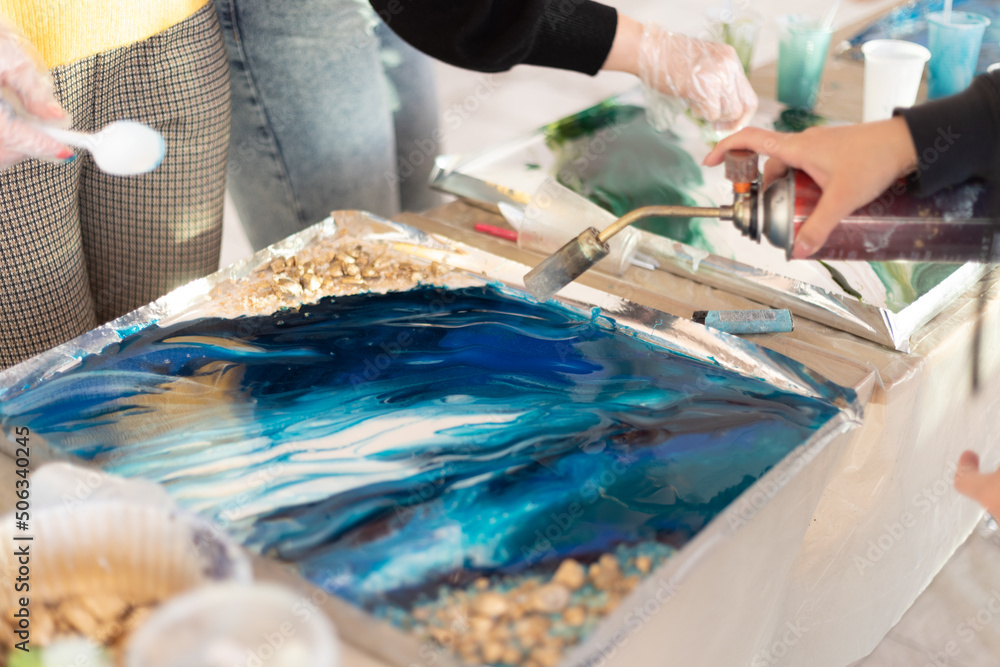 Drying paint in blue color with a hairdryer on the board. Female hand holds a manual gas burner, create a painting in resin art technique