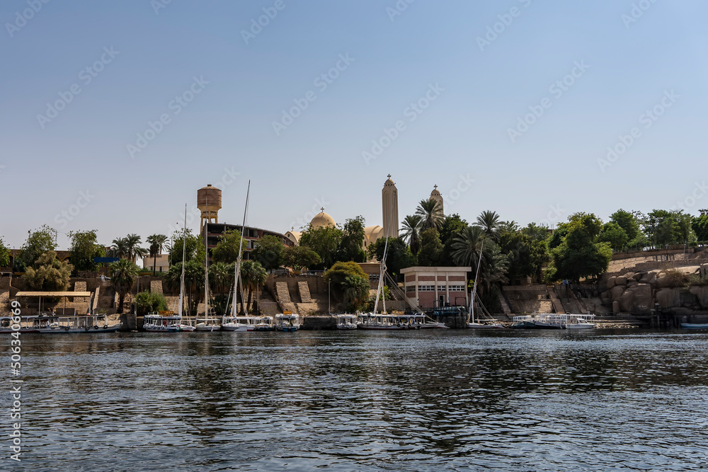Tourist boats and felucca moored at the river bank. City houses are visible among the green vegetation. Ripples and reflections on the river. Egypt. Nile