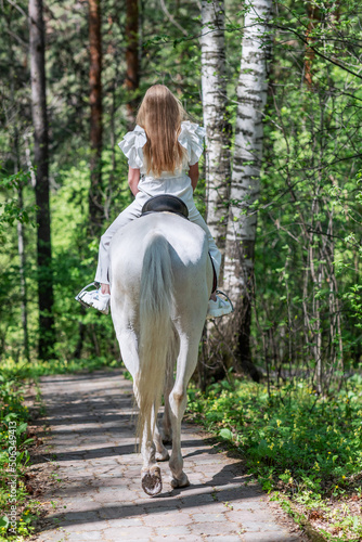 The girl rides a white horse with her back to the viewer in the spring forest. Communication with animals in nature. 