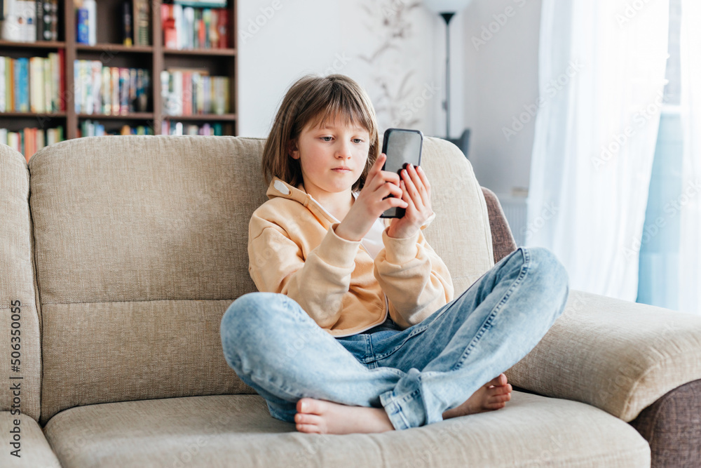 Schoolgirl is sitting on the sofa and playing mobile phone or chatting with friends. Children use technology. Modern gadgets and teenagers. Schoolgirl is sitting on the sofa and playing mobile phone or chatting with friends. Children use technology. Modern gadgets and teenagers.
