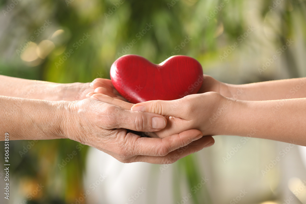 Young and elderly women holding red heart in hands on blurred green background, closeup Young and elderly women holding red heart in hands on blurred green background, closeup